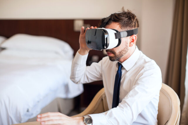 Mann sitzt auf Bett im Hotelzimmer und hat VR-Brille auf