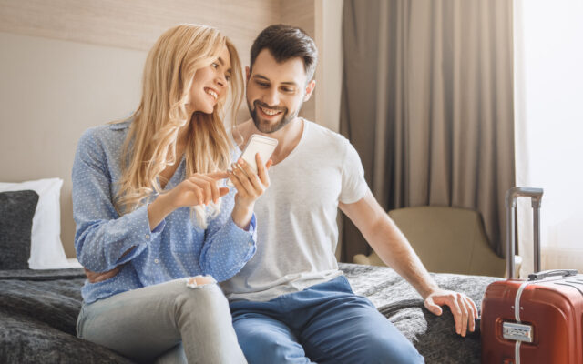 Lächelndes Paar sitzt im Hotelzimmer auf dem Bett und schaut auf ein Smartphone