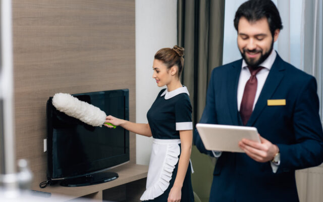 Glücklicher Hoteldirektor mit Tablet und reinigendes Zimmermädchen im Hotelzimmer