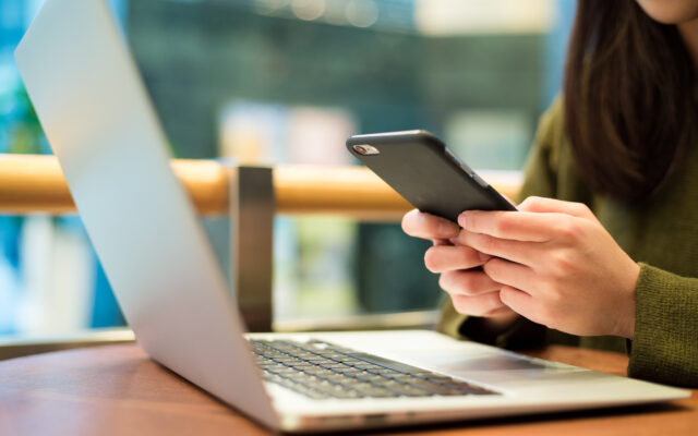 Eine Frau sitzt vor dem Laptop und tippt auf Smartphone