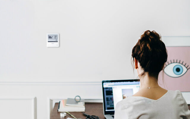 Frau sitzt am Schreibtisch im Büro und schaut auf Wand mit fan coil Thermostat
